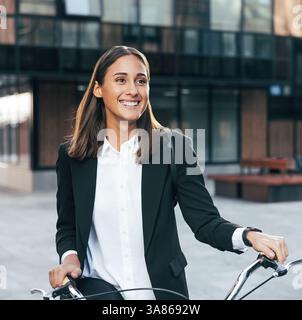 Felice imprenditrice donna in piedi all'aperto con la bicicletta in un edificio aziendale. Ritratto di una donna d'affari sorridente che indossa un abbigliamento formale e uno zaino Foto Stock