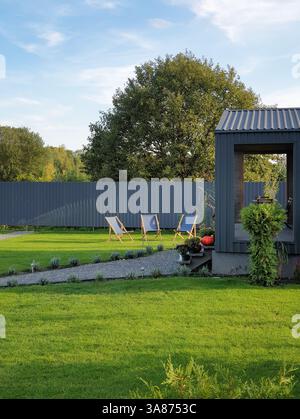 Cortile posteriore con i raggi del sole pomeridiano: Tre sdraio su un prato, portico della casa estiva e sentiero pedonale. diversi alberi dietro la recinzione grigia. Foto Stock