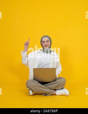 La ragazza musulmana caucasica punta sopra. La ragazza indossa abaya hijab seduto sul pavimento del computer portatile in grembo. Ho una grande idea idea idea idea immagine. Giallo. Foto Stock