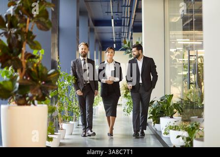 Professionisti aziendali sicuri che camminano attraverso i corridoi degli uffici con piante verdi, discutendo del lavoro. Foto Stock