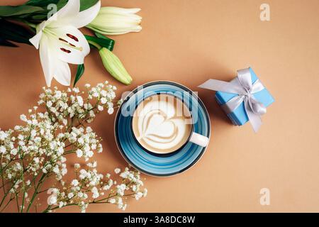 Tazza da caffè con cuore latte art, gigli bianchi, scatola regalo e fiori su sfondo beige. Vista dall'alto, disposizione piatta. Foto Stock
