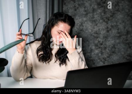 Sintomi di eccessivo lavoro. Giovane, stanca e libera professionista con gli occhiali seduti davanti al computer che sfrega gli occhi sentendo affaticamento degli occhi durante il lavoro. Foto Stock