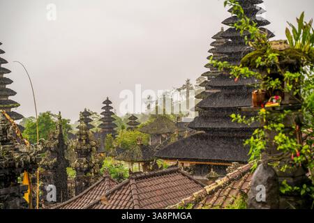 La pioggia nebbiosa cade sull'antico complesso di templi balinesi con caratteristici tetti a pagoda a più livelli annidati nel verde lussureggiante, creando un'atmosfera mistica Foto Stock
