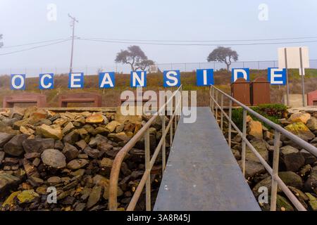 Oceanside, California - 3 gennaio 2025: Vista chiara del cartello Oceanside con primo piano roccioso e sentiero costiero, scattato durante una mattinata nebbiosa a O. Foto Stock