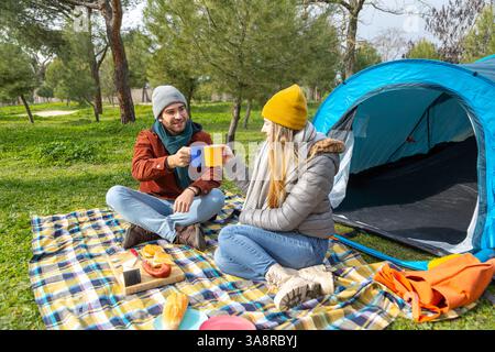 Giovani coppie che si godono un picnic fuori dalla loro tenda, brindando con tazze da caffè in un bellissimo parco Foto Stock