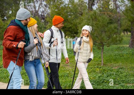 Quattro amici si stanno godendo un'escursione in un parco durante l'inverno, utilizzando bastoncini da trekking e zaini, parlando e sorridendo Foto Stock