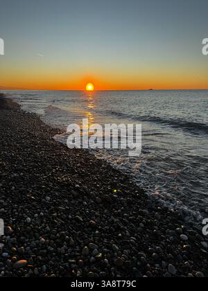 Un tramonto dorato su una spiaggia di ciottoli, con onde delicate che lavano a riva e riflessi scintillanti sull'acqua. Foto Stock