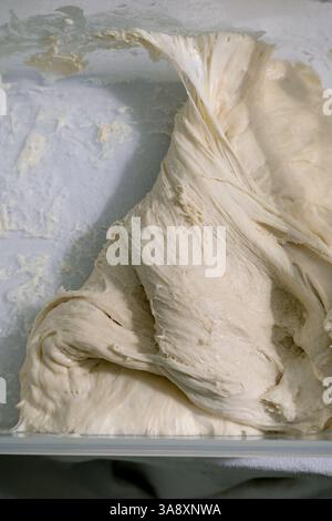 Vista ravvicinata della consistenza cremosa dell'impasto piegato durante la preparazione della cottura. Impasto bianco elastico con superficie liscia e lucida pronto per l'impasto. Foto Stock