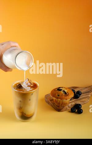 Una persona sta versando del latte in una tazza di caffè. Il caffè viene servito su un tavolo giallo con un muffin e mirtilli sul bordo di legno. Foto Stock