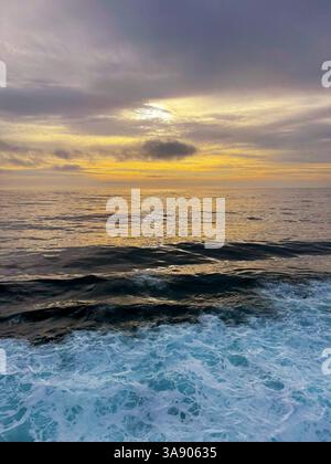 Il sole tramonta sull'oceano aperto, gettando luce dorata sulle onde dolci e facendo surf schiumoso sotto un cielo spettacolare pieno di nuvole a strati. Foto Stock