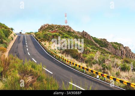 La strada tortuosa con barriere di sicurezza a righe curva attraverso una vegetazione lussureggiante, che conduce alla cima di una collina. La ripida strada di montagna si snoda verso l'alto. Carreggiata asfaltata Foto Stock