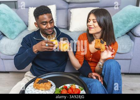 Una coppia multietnica si siede insieme, gustando la colazione e il vino, creando un'esperienza culinaria intima e intima nel loro soggiorno Foto Stock