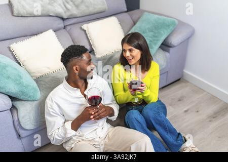 Coppia gioiosa seduta sul pavimento, sorseggiando vino e condividendo un momento di connessione in un accogliente soggiorno con morbidi cuscini Foto Stock