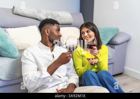 La coppia si diverte un momento insieme, condividendo vino e sorrisi mentre si siede comodamente a casa, creando un'atmosfera calda e intima Foto Stock