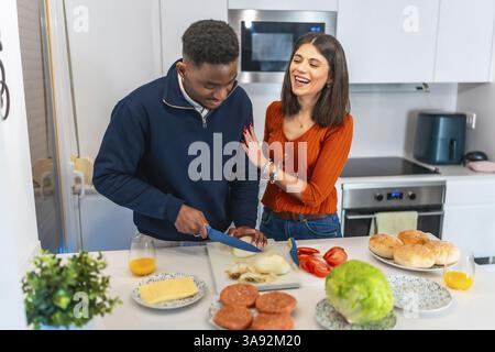 Allegra coppia multietnica che si diverte mentre preparano hamburger per cena nella loro cucina moderna Foto Stock