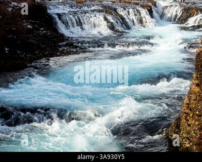 Cascata Bruarfoss, Islanda: Cascate di acque turchesi cristalline sopra l'aspra roccia vulcanica, che creano un contrasto sorprendente tra blu ghiaccio e scuro Foto Stock
