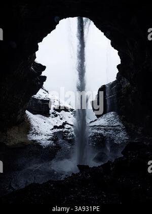 La cascata di Kvernufoss vista da dietro, incorniciata da un'apertura scura della grotta. L'acqua a cascata contrasta nettamente con la neve e la luce esterna, creato Foto Stock