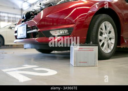 Primo piano di una batteria di un'auto sul pavimento di un'officina accanto a un veicolo rosso, raffigurante la manutenzione e la sostituzione della batteria. Foto Stock