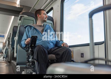 Uomo in sedia a rotelle che viaggia in treno, con una valigia, godendo di un paesaggio attraverso il finestrino del veicolo in movimento. Foto Stock
