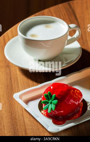 Mousse cake con ripieno di fragole ricoperto di glassa a specchio e una tazza di latte su un tavolo in un caffè. Dessert, bevande, tavolo di legno in un caffè cittadino, se Foto Stock