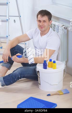 Un bell'uomo che indossa abiti da lavoro seduto sul pavimento regge il paintroller e guarda la macchina fotografica Foto Stock