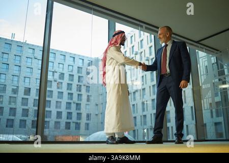 Grandangolo basso di due diversi partner commerciali maschi che si salutano con una stretta di mano mentre si trovano in un ufficio spazioso con finestre panoramiche Foto Stock