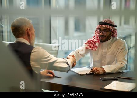 Attraverso una parete di vetro di due partner commerciali etnie diverse che stringono la mano quando fanno un affare Foto Stock