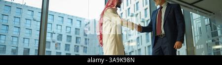 Grandangolo basso di due diversi partner commerciali maschi che si salutano l'un l'altro con una stretta di mano mentre si trovano in un ufficio spazioso con finestre panoramiche, Foto Stock
