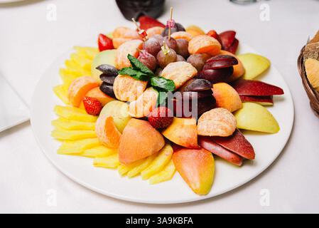 Frutta assortita a fette, tra cui ananas, mele, mandarini, uva, fragole, e prugne servite su un piatto bianco per un pasto festivo. Foto Stock