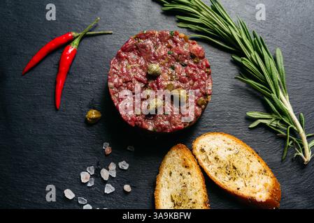 Vista dall'alto della tartare di manzo tagliata finemente con capperi, rosmarino, peperoncino e crostini alle erbe su ardesia scura. Foto Stock