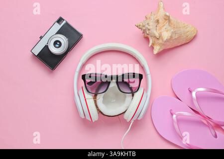 Creativi scenari estivi sulla spiaggia. Vacanze al mare. Cuffie, occhiali da sole, noce di cocco, guscio, infradito, fotocamera retrò su sfondo rosa pastello. Vista dall'alto. Foto Stock