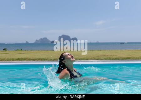 La donna si rilassa in una scintillante piscina, spruzzando acqua gioiosamente mentre si prende il sole. Le splendide montagne si innalzano sullo sfondo, creando un'atmosfera mozzafiato Foto Stock