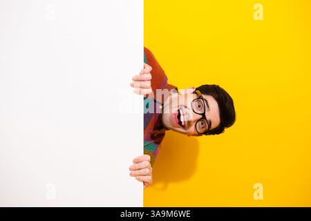 Giovane eccitato che sbircia da dietro una lavagna bianca, sorridendo con entusiasmo su uno sfondo giallo brillante Foto Stock