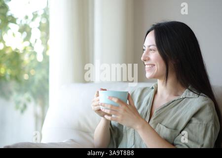 Felice donna asiatica che beve caffè e guarda attraverso una finestra seduta su un divano a casa Foto Stock