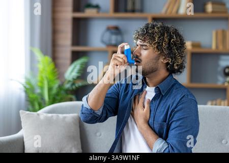 Un giovane con i capelli ricci sta usando un inalatore per l'asma mentre si siede su un divano in un luminoso salotto. Foto Stock