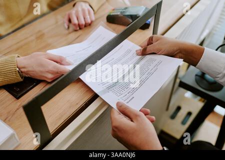 Mani di due persone che firmano un documento ufficiale presso uno sportello di un ufficio, con un terminale di pagamento e altri materiali in background, atmosfera collaborativa per le trattative d'affari Foto Stock