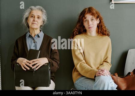 Ritratto di una donna anziana seduta accanto a una giovane donna sul divano, che guarda la macchina fotografica, mostra legami generazionali e condivide espressioni ponderate Foto Stock