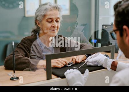 Donna anziana che esamina attentamente i gioielli al bancone con l'aiuto di un gioielliere, riflettendo una considerazione premurosa Foto Stock