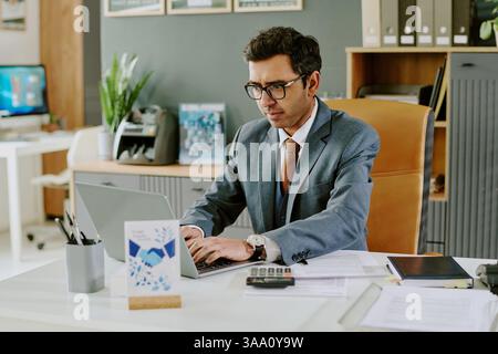 Uomo d'affari ispanico che indossa una tuta che lavora su un computer portatile mentre è seduto alla scrivania in un ufficio moderno, concentrandosi sulle attività con documenti e organizzatori visibili sul tavolo Foto Stock