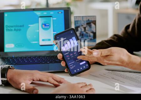 Due colleghi analizzano i dati su uno smartphone mentre sono seduti alla scrivania dell'ufficio, collaborano al progetto, sul laptop vengono visualizzati app mobili e documenti visibili Foto Stock