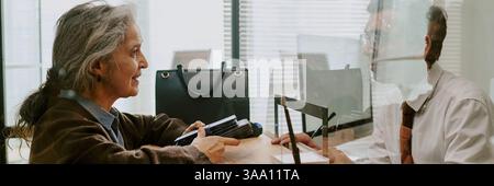 Donna anziana con capelli grigi impegnata in incontri d'affari con uomo in abbigliamento formale attraverso una partizione in vetro in un moderno ambiente da ufficio Foto Stock
