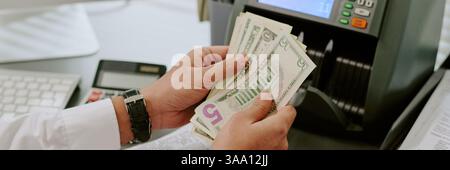 Mano che tiene in mano più dollari mentre si utilizza il bancomat alla scrivania con tastiera e calcolatrice visibili in background Foto Stock