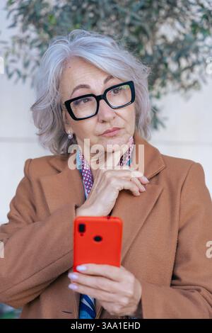 Donna anziana premurosa che utilizza lo smartphone all'aperto in ambienti urbani.verticale Foto Stock