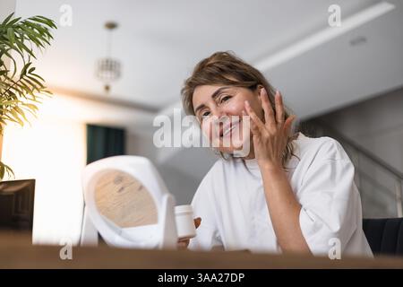 Una donna sorride mentre applica un prodotto per la cura della pelle al viso in uno spazio interno ben illuminato. La luce naturale esalta la sua espressione gioiosa durante il suo ingresso Foto Stock