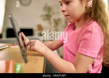 Ragazza concentrata in camicia rosa che interagisce con il tablet durante la sessione di apprendimento a distanza a casa, utilizzando con attenzione lo schermo tattile. Foto Stock