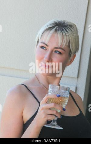 bella donna adulta che beve vino bianco mentre si trova accanto alla finestra Foto Stock