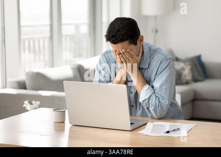 L'uomo copre il viso con le mani sedute alla scrivania con un computer portatile Foto Stock