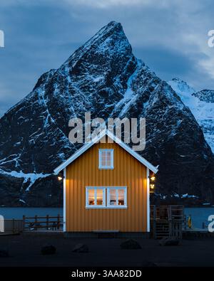 Winter auf den Lofoten in Norwegen mit traditionell gelbem Rorbua in Sakrisoy und verschneitem Olstinden im Hintergrund zur blauen Stunde am Fjord Foto Stock