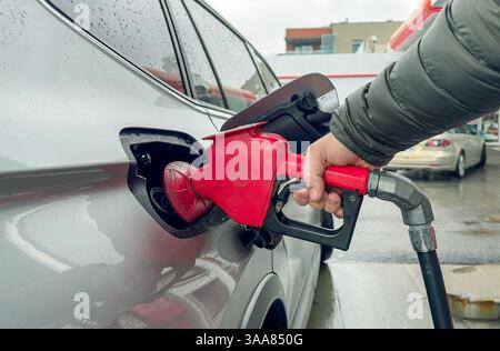 Primo piano di una persona che fa rifornimento di un'auto presso una stazione di servizio, con un ugello rosso inserito nel serbatoio. Foto Stock
