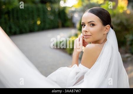 Splendida sposa in posa graziosamente con un velo bianco che scorre, irradia gioia ed eleganza il giorno del suo matrimonio in un tranquillo parco Foto Stock